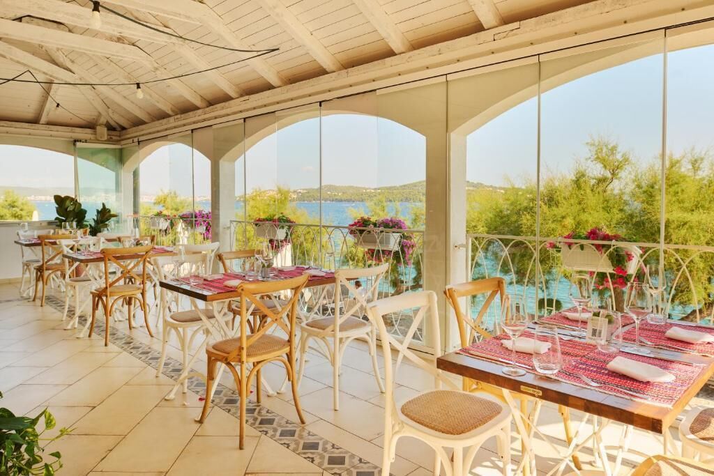 Un'area pranzo esterna riparata, con tavoli e sedie, ampie finestre e vista sul mare.