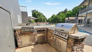 An outdoor kitchen with a grill, sink, and storage cabinets, adjacent to a swimming pool and a covered seating area. - Green & Private 2BR King Bed/Pool/Gym Near UAB/Hospitals (Homewood)
