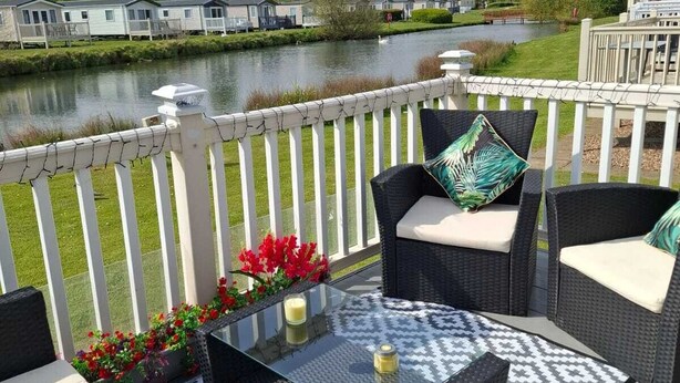 Een terras met rieten meubels, een glazen tafel en uitzicht op een meer en stacaravans op de achtergrond.