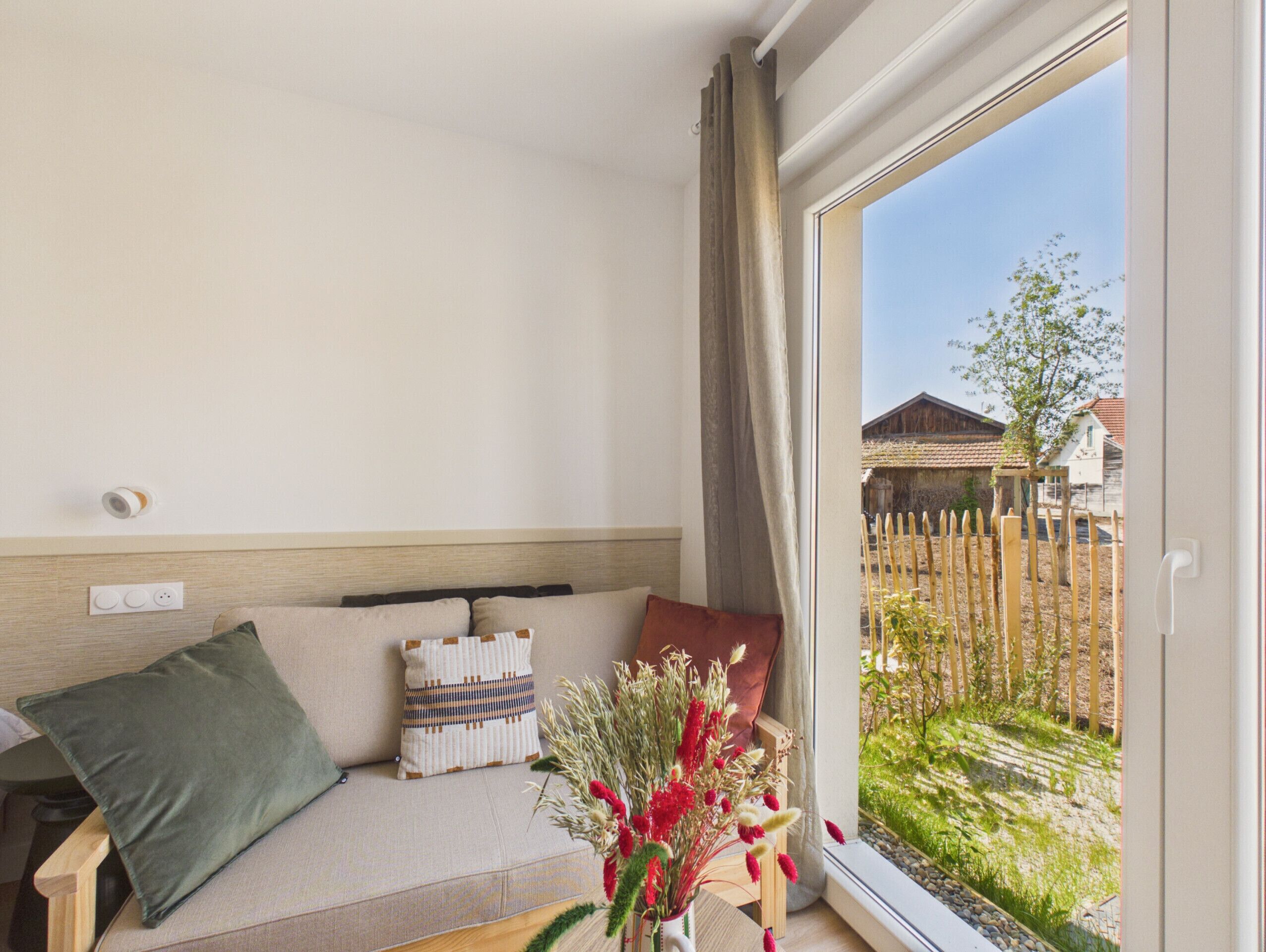A room with a sofa, a vase with flowers, a window with a view of a fence and buildings.