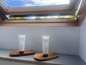 Une fenêtre de toit laissant entrer la lumière naturelle, avec deux tubes de gel douche et d’après-shampoing de la marque ESPA, posés sur des supports en bois.