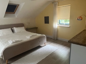 Une chambre à coucher comprenant un lit, une table de chevet, une commode, une fenêtre avec des rideaux, un luminaire au plafond et une petite lucarne.