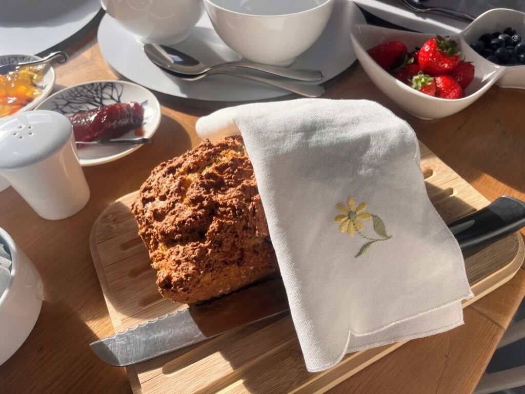 Une table mise pour le petit-déjeuner avec un muffin sur une planche en bois, une nappe blanche à motifs floraux, un bol de fraises et une tasse de café.