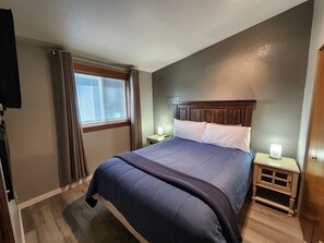 Ein Schlafzimmer mit einem Bett, zwei Nachttischen, einem Fenster mit Vorhängen, einem Wand-Fernseher und einem Beistelltisch mit Lampe.