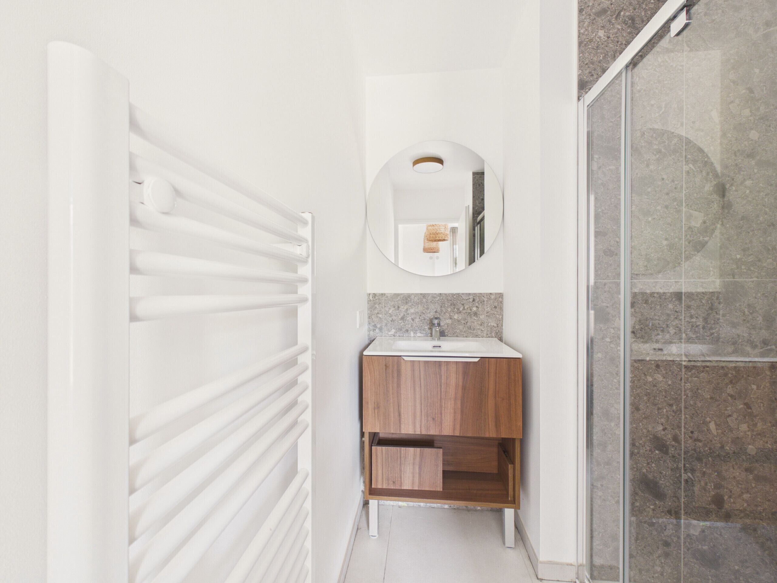 Een badkamer met een witte radiator, een ronde spiegel, een houten wastafelmeubel en een douchecabine van glas.