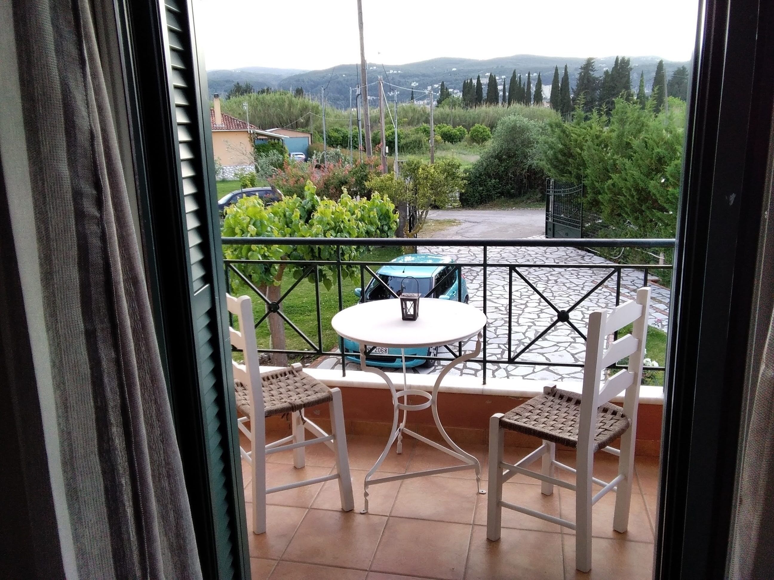 A balcony with a table and chairs, a view of a garden, and a car parked outside.