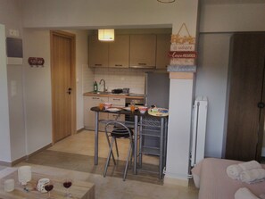 A compact kitchen with a sink, a small dining table, and a chair. There is a refrigerator, a cabinet, and a wall-mounted shelf. - Casa memories (Kalligoni)