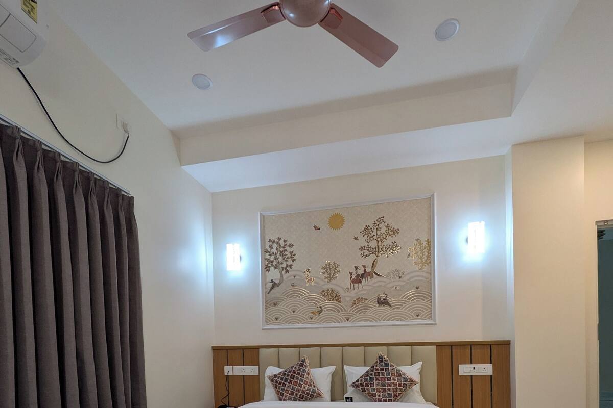 Una habitación de hotel con cama, ventilador de techo, una obra de arte en la pared y ventana con cortinas.