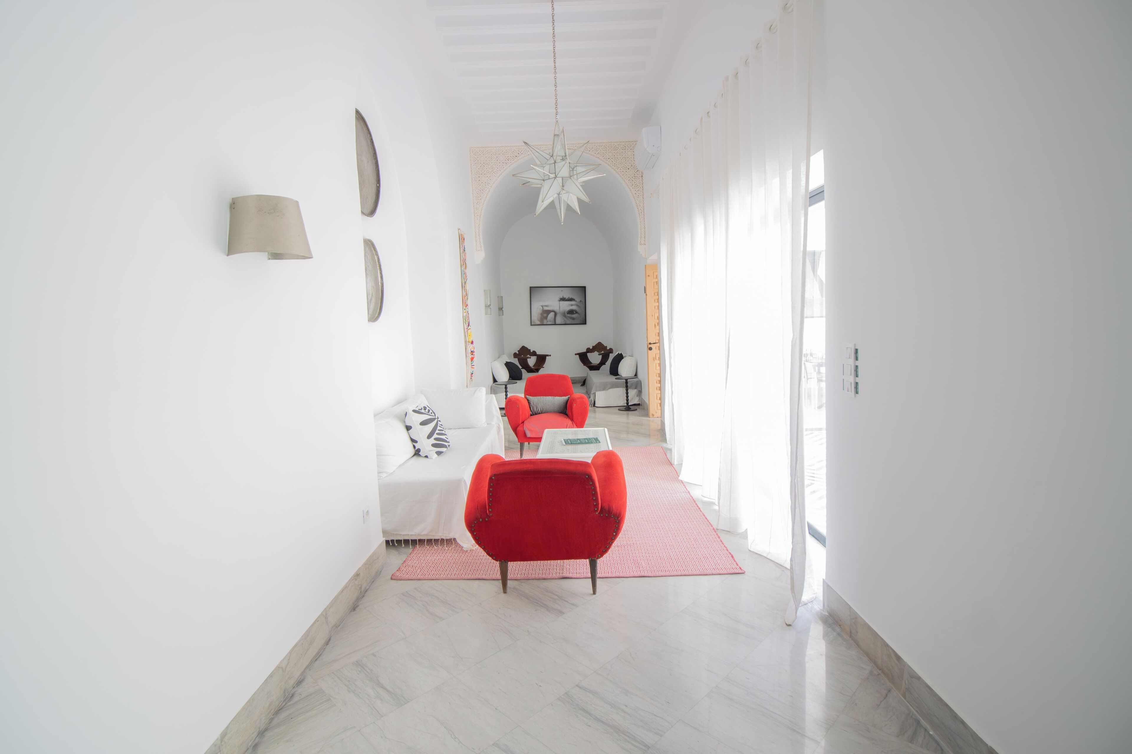 Un couloir mène à un espace de vie comprenant un fauteuil rouge, un canapé blanc et un luminaire suspendu au plafond.