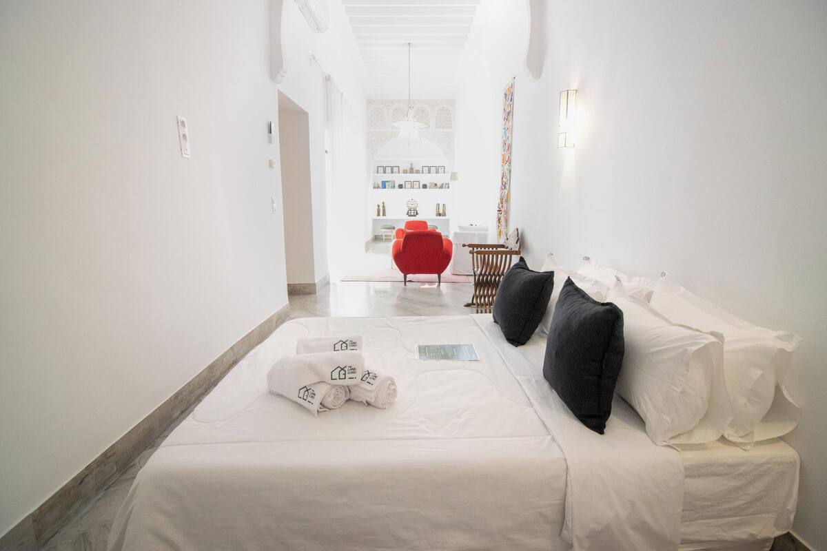 A hotel room with a bed, a red chair, a wooden bench, and a white wall with a light fixture.