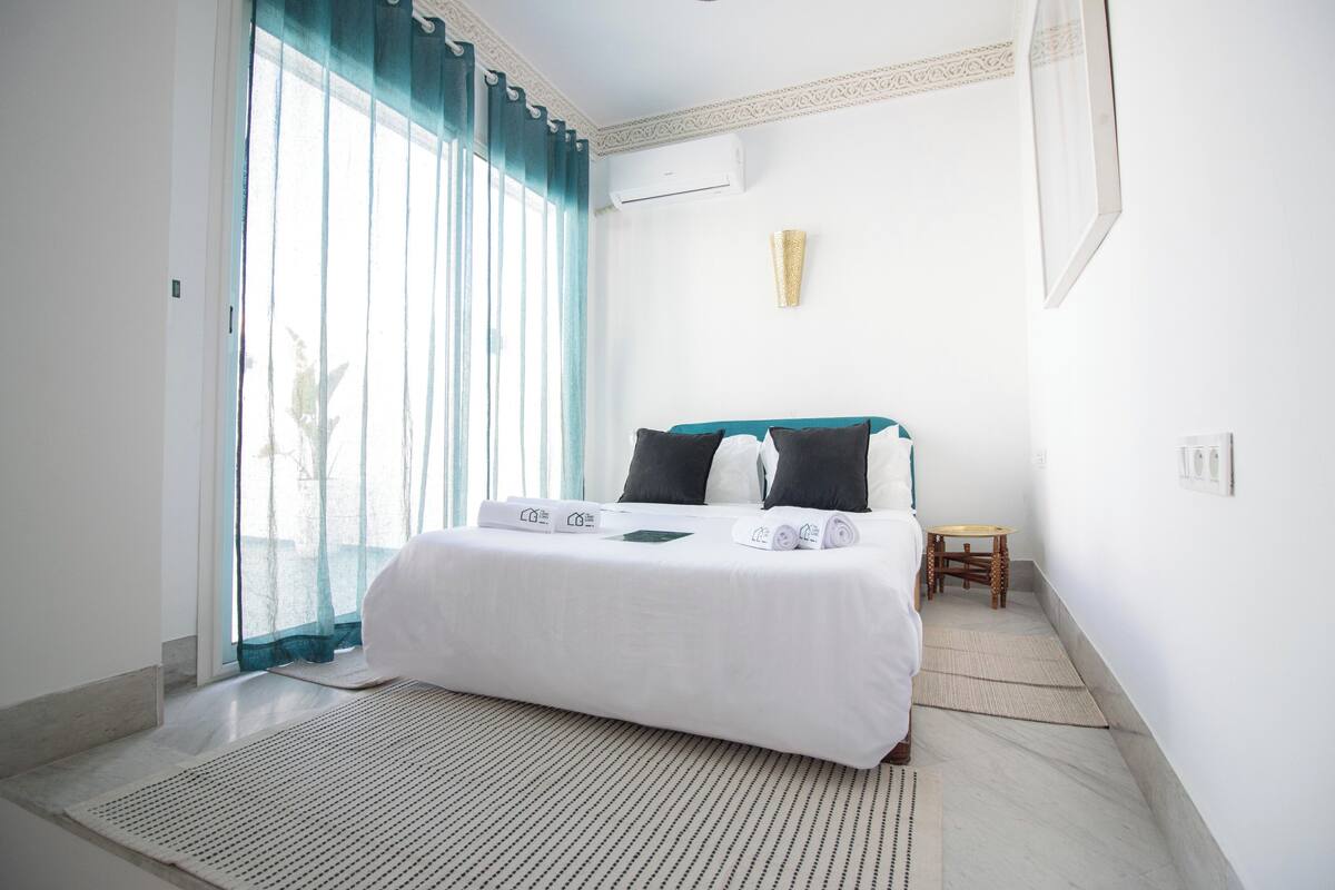 A hotel room with a bed, a small table, a wall-mounted air conditioner, and a window with curtains.
