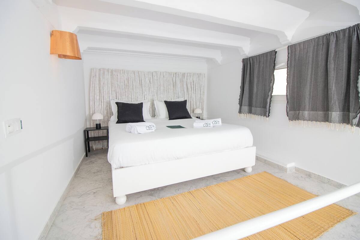 A bedroom with a white bed, a nightstand, a lamp, and a window with curtains.