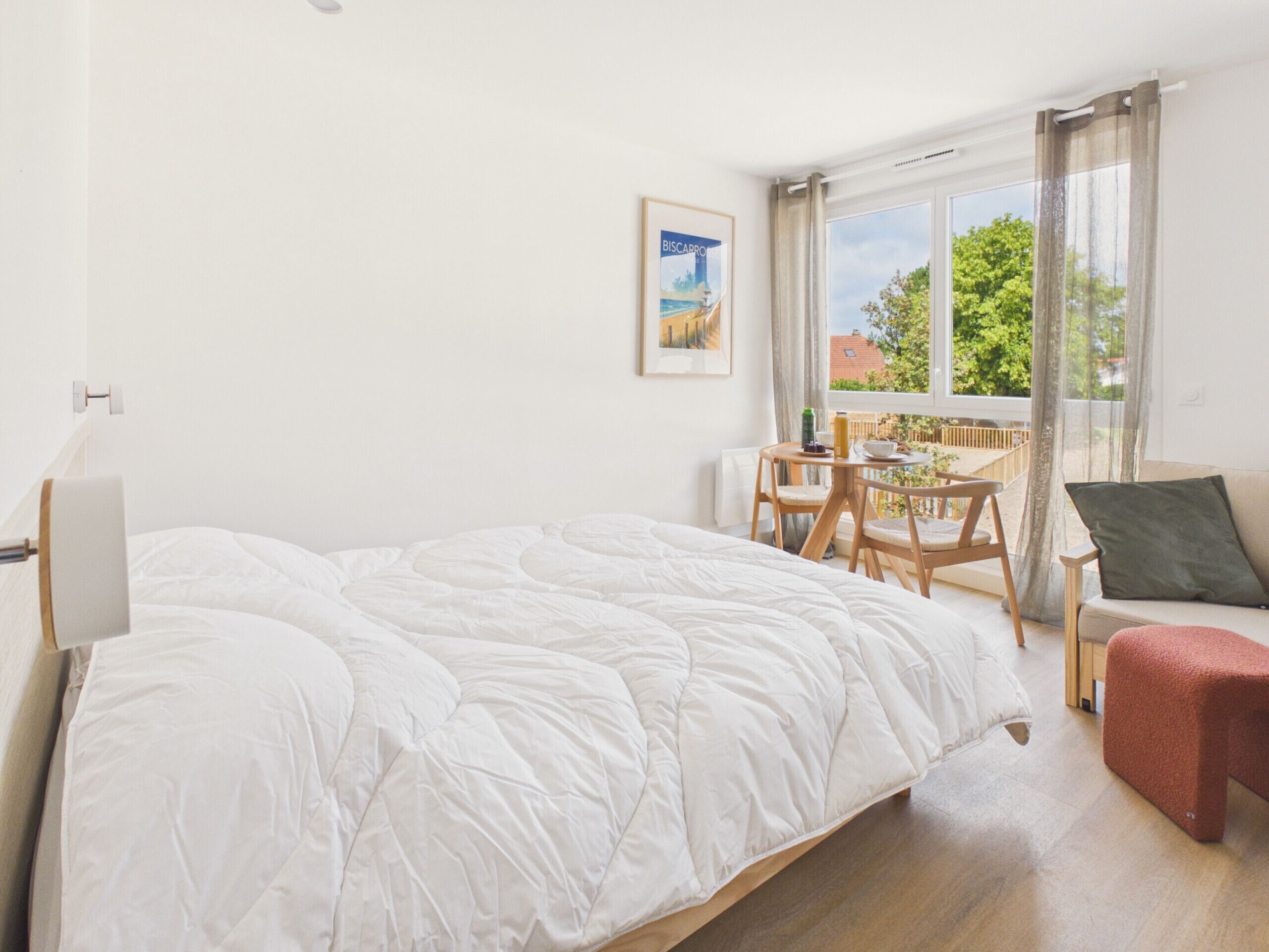 A bedroom with a bed, a small table with chairs, a sofa, and a window with a view of greenery.