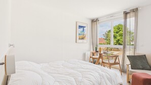 A bedroom with a bed, a small table with chairs, a sofa, and a window with a view of greenery. - Le Curl - studio for 3 people - shared pool (Biscarrosse)