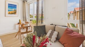 A modern living room with a sofa, a small table with a bottle and a bowl, a chair, and a window with a view of houses. - Le Curl - studio for 3 people - shared pool (Biscarrosse)