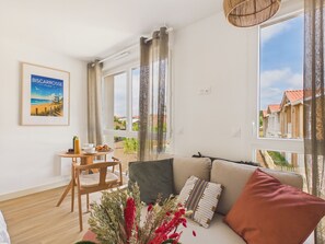 A modern living room with a sofa, a small table with a bottle and a bowl, a chair, and a window with a view of houses. - Le Curl - studio for 3 people - shared pool (Biscarrosse)