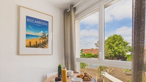 A dining area with a table set for two, a framed picture of a beach scene, and a view of a residential area through a window. - Le Curl - studio for 3 people - shared pool (Biscarrosse)