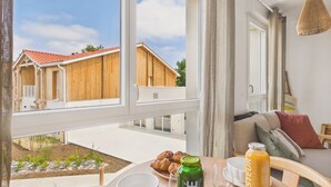 A room with a view of wooden buildings, a table with croissants, a bottle of juice, and a sofa with cushions. - Le Curl - studio for 3 people - shared pool (Biscarrosse)