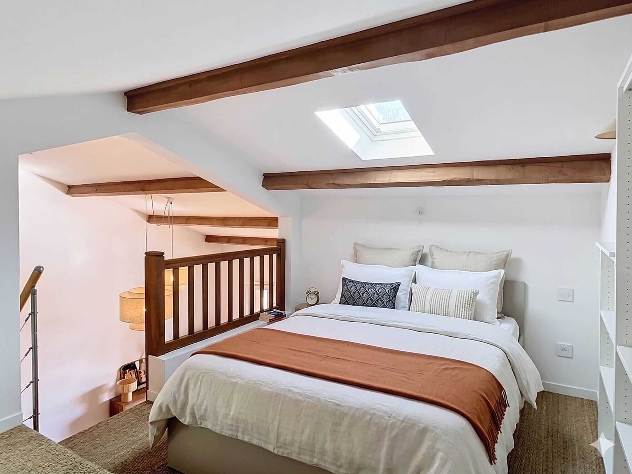 A bedroom with a sloped ceiling, a skylight, a bed with multiple pillows, a bedside table, and a lamp.