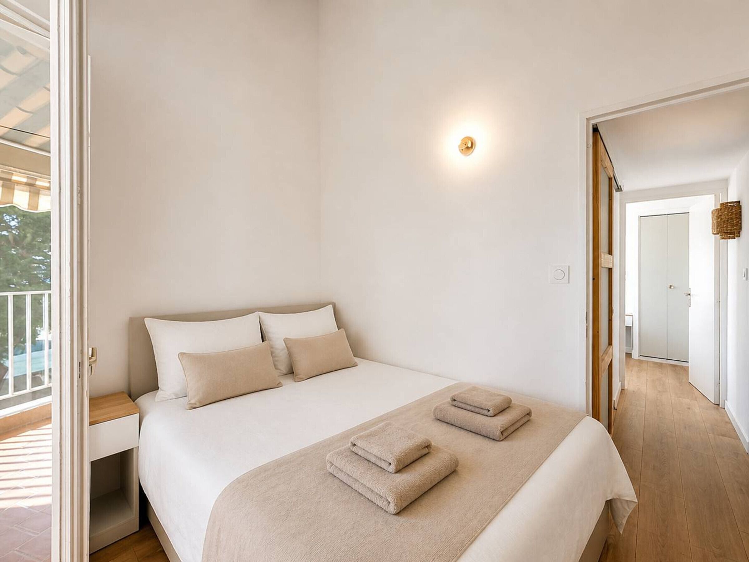 A bedroom with a bed, pillows, a bedside table, a wall-mounted light, and a view of the outside.