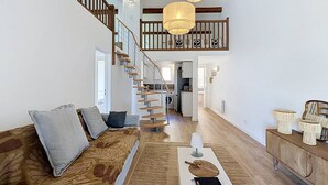 A spacious living room with a wooden staircase, a sofa with cushions, a coffee table with a book and a lamp, and a dining area with a wooden cabinet and lamps. - Apartment near beach with parking - 4 guests (Saint-Cyr-sur-Mer)