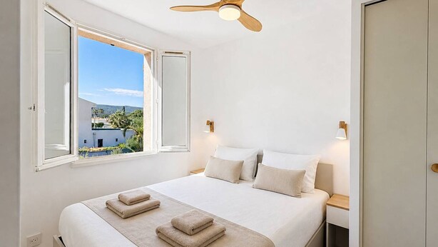 A bedroom with a bed, a ceiling fan, a window with an open shutter, and a view of a building and trees. - Apartment near beach with parking - 4 guests (Saint-Cyr-sur-Mer)
