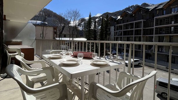 A balcony with a table set for four, surrounded by white plastic chairs, overlooking a mountainous area with buildings in the background. - Spacious apartment with balcony - 4 guests (Valloire)