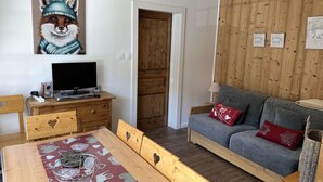 A cozy living room with a wooden dining table, a grey sofa with red cushions, a television on a wooden stand, and a wall with a framed animal painting. - Spacious apartment with balcony - 4 guests (Valloire)