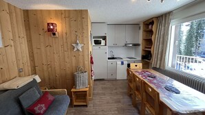 A cozy room with a wooden wall, a sofa, a dining table, and a kitchen area with cabinets and a microwave. - Spacious apartment with balcony - 4 guests (Valloire)