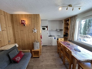 A cozy room with a wooden wall, a sofa, a dining table, and a kitchen area with cabinets and a microwave. - Spacious apartment with balcony - 4 guests (Valloire)
