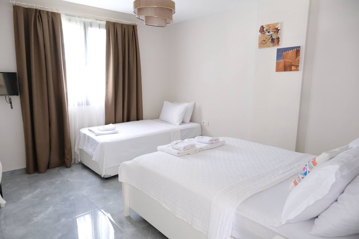 A hotel room with two beds, a flat-screen TV, and a window with curtains.