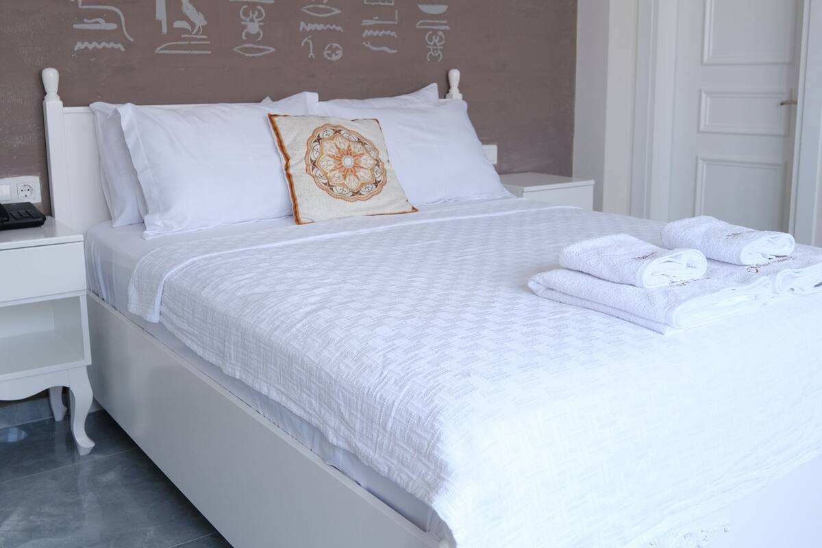 A white bed with a patterned bedspread, a decorative pillow, and neatly folded towels.