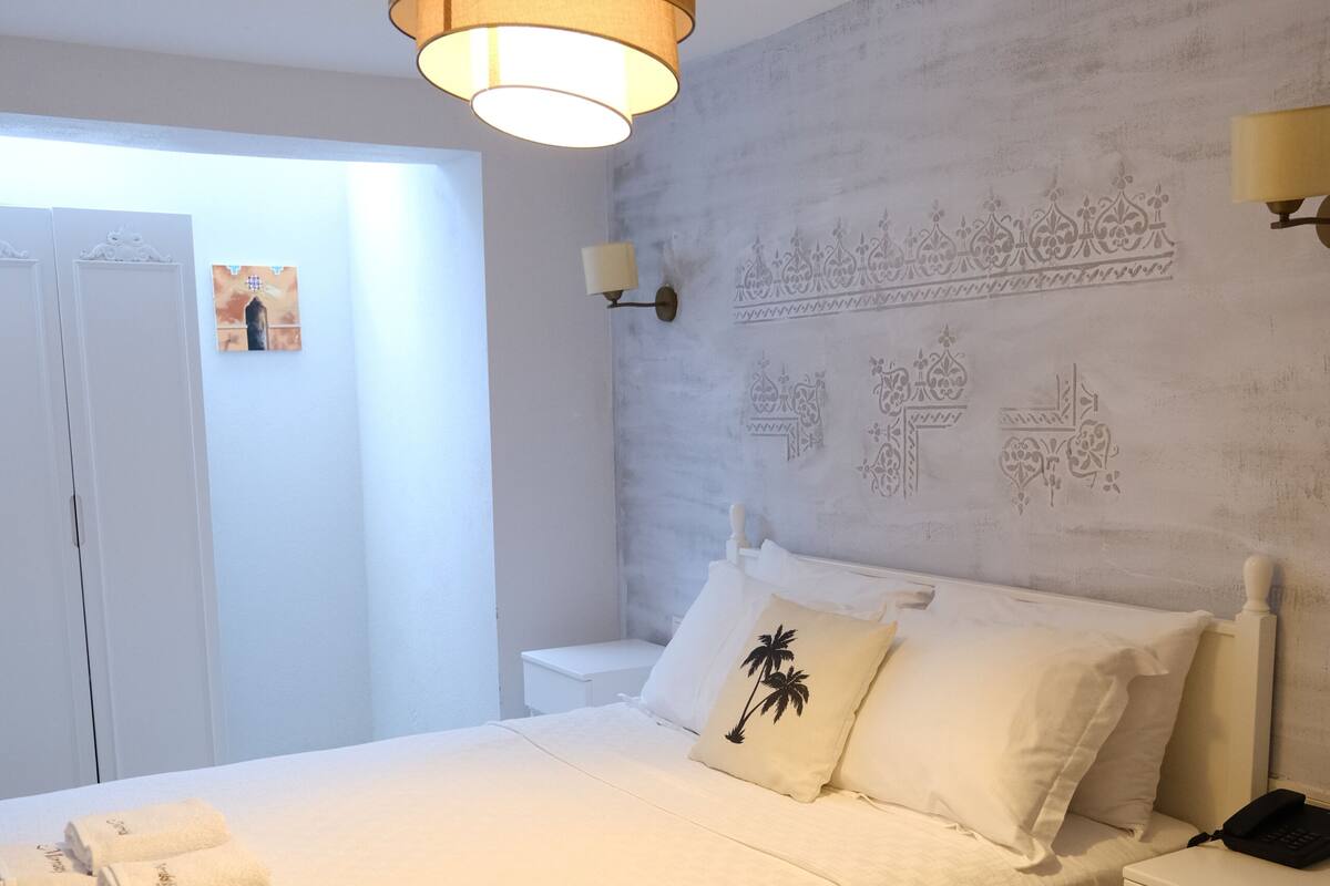 A white bed with a headboard, bedside tables, a hanging light fixture, and a wall with decorative patterns.