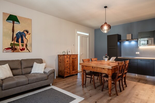 Ein Wohnzimmer mit einer Couch, einem Holzesstisch und Stühlen. Es gibt eine Küchenzeile mit Schränken und einer Mikrowelle.