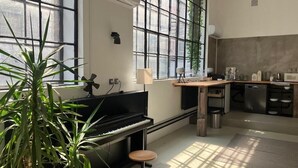 Quarto com piano, mesa de jantar, cozinha e janelas amplas.