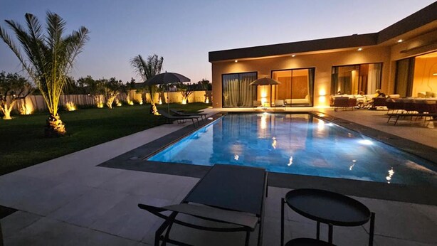 Una piscina con sillas de descanso, un patio y una casa moderna con ventanas amplias.