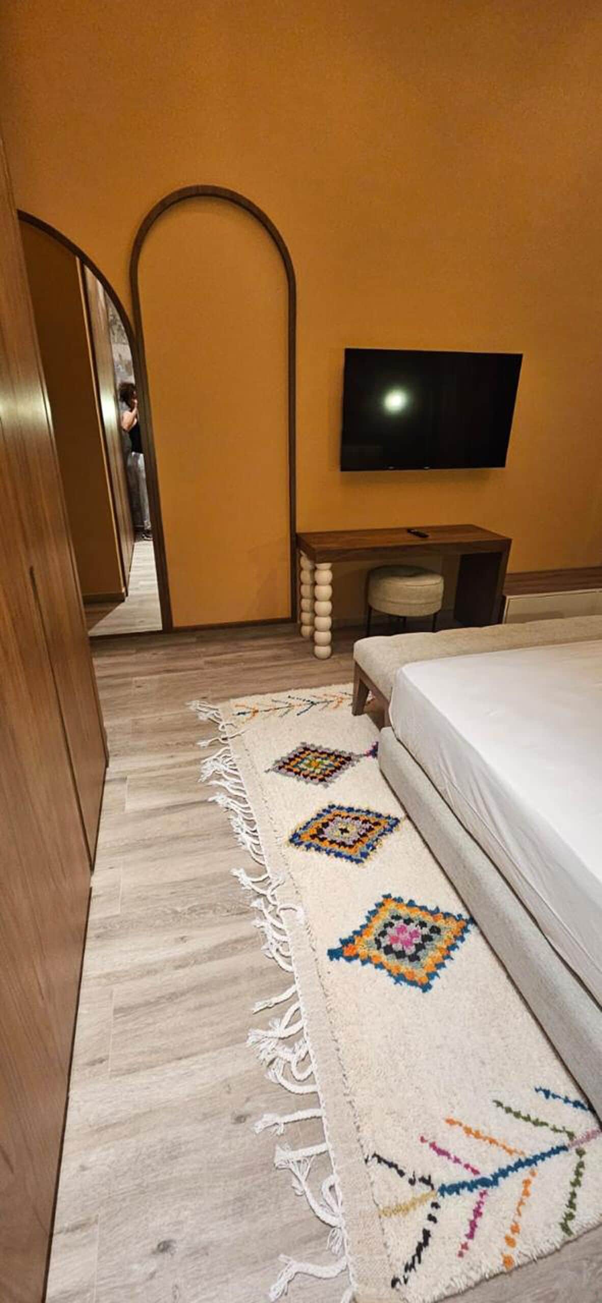 A hotel room with a bed, a TV mounted on the wall, a small table with a stool, and a patterned rug.