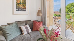 A living room with a sofa, pillows, a framed picture, a lamp, and a view of the outdoors. - L'Ondée - apartment for 4 people - shared pool (Biscarrosse)