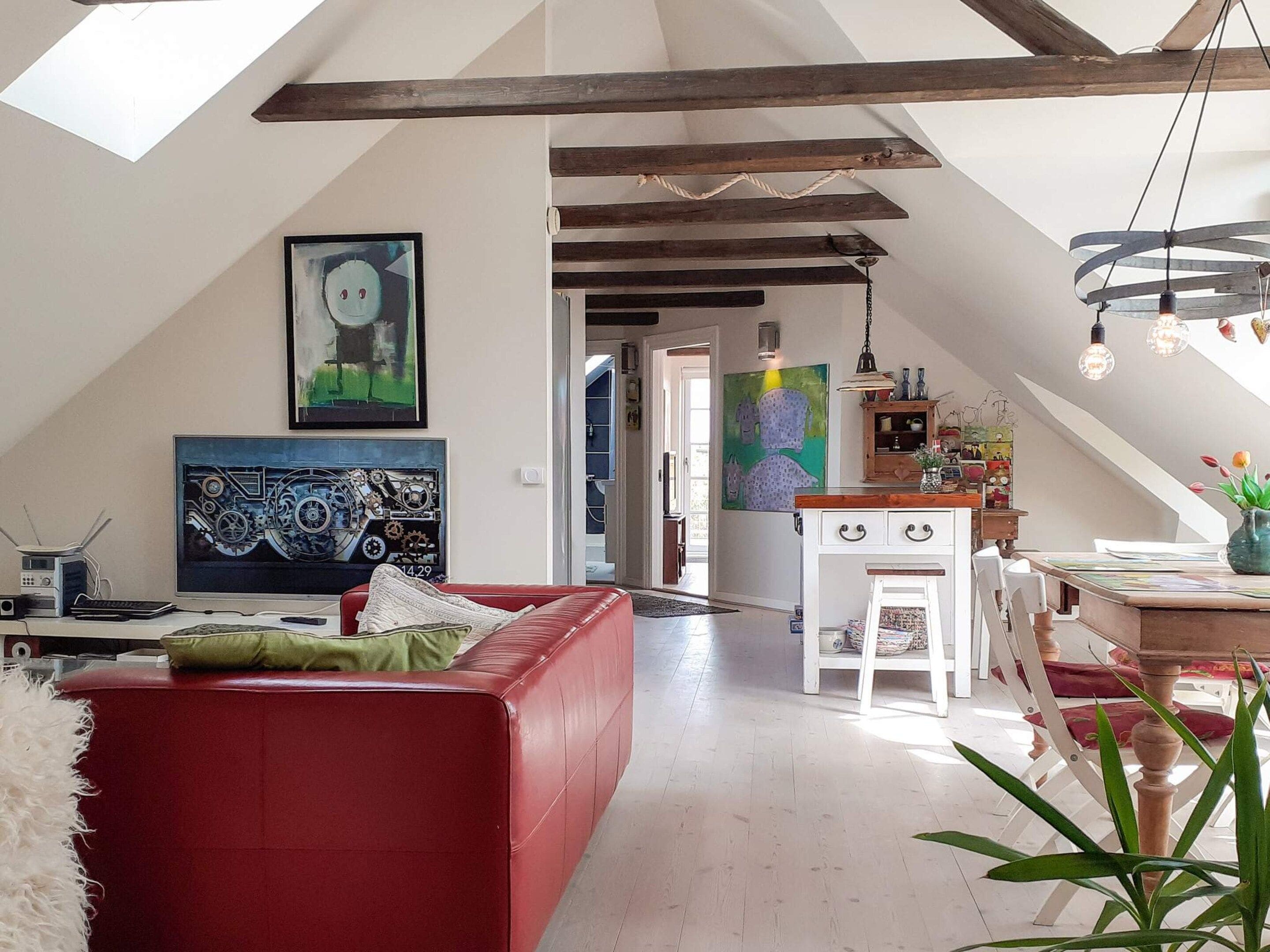A spacious loft with a high ceiling, exposed beams, a red leather sofa, a dining area with a wooden table, and artwork on the walls.