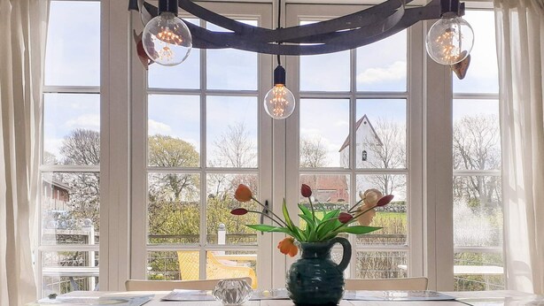 A dining area with a table set for two, a vase with flowers, and a view of a house through large windows. - 7 Person Holiday Home in Logstrup (Logstrup)