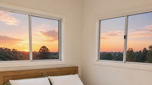 A bedroom with a wooden headboard, two windows, a bed with white linens, and a view of a sunset.