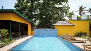 Una piscina con borde de azulejos, rodeada de un patio con sillas y una pequeña construcción con techo de baldosas.