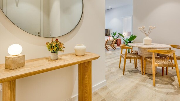 A modern interior with a round mirror, a wooden console table with a vase and a light, wicker chairs around a small round table, and a view into a living area with a plant and a sofa. - Modern apartment near the marina - Estepona (Estepona)
