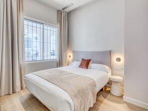 Un dormitori modern amb un llit gran, una taula de nit, una tauleta auxiliar i una finestra amb cortines, més una làmpada penjada a la paret.
