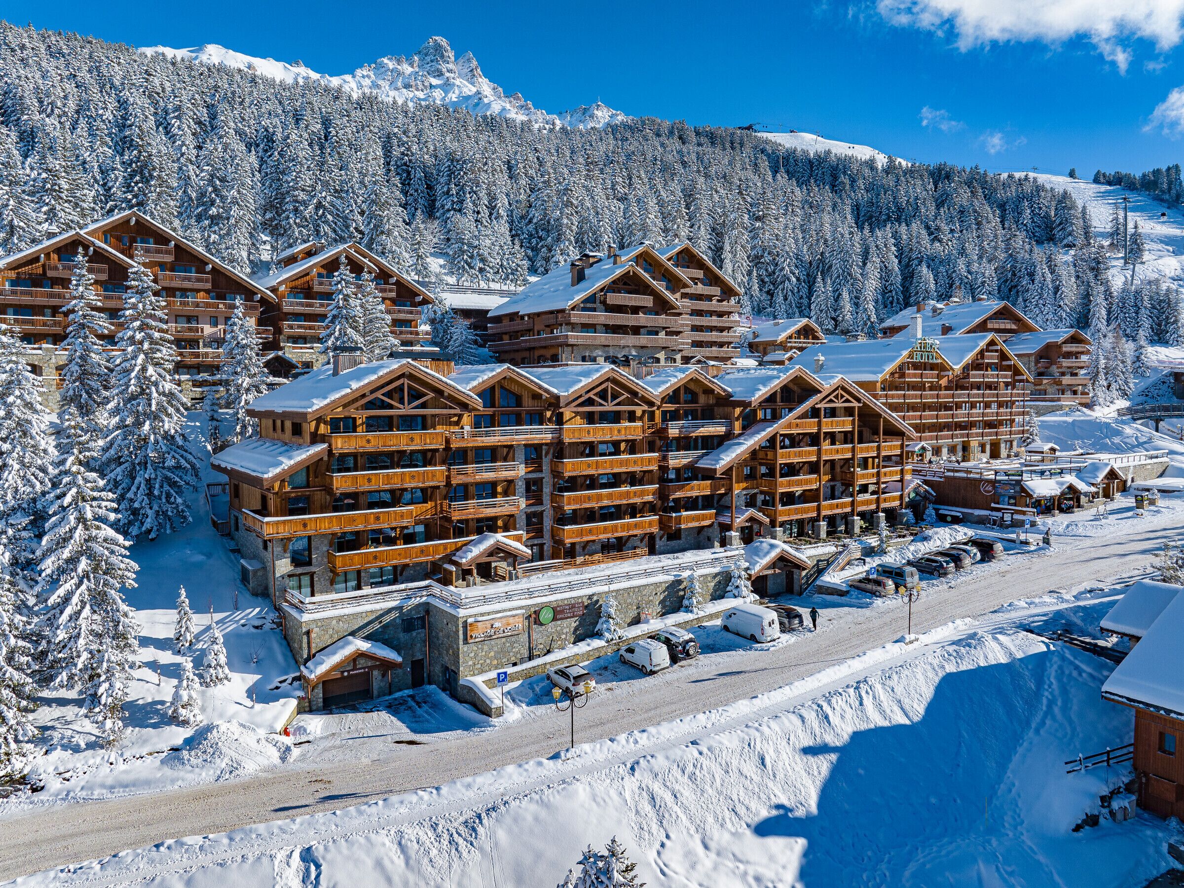Un chalet enneigé, composé de plusieurs bâtiments en bois, avec une piste de ski en arrière-plan et un ciel dégagé.