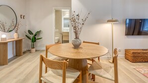 Ett modernt matområde med ett runt träbord, fyra stolar, en vas med torkade växter, en golvlampa och en tv som är monterad på väggen.