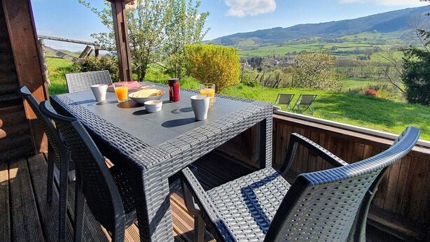 An outdoor dining area with a table set for two, surrounded by wicker chairs, overlooking a scenic landscape with hills and a clear sky.