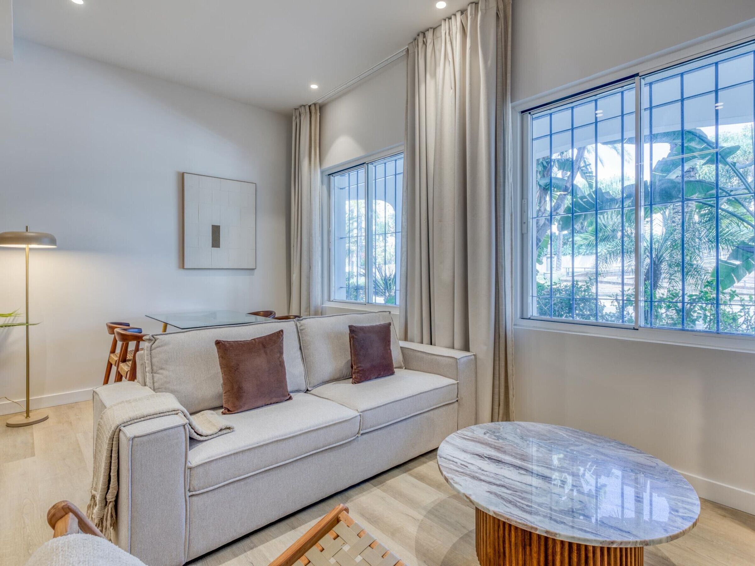 Uma sala de estar moderna com um sofá, uma mesa de centro redonda e grandes janelas com cortinas.