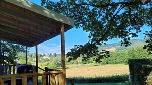 A wooden deck with chairs, a covered area, and a view of a green field and hills. - Mobile home with air conditioning - 6 guests (Saint-Point)