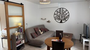 A modern living room with a sofa, a dining table, a bookshelf, a decorative tree wall art, and a pendant light. - Peaceful cottage, parking, pets allowed (Saint-Georges-sur-Cher)